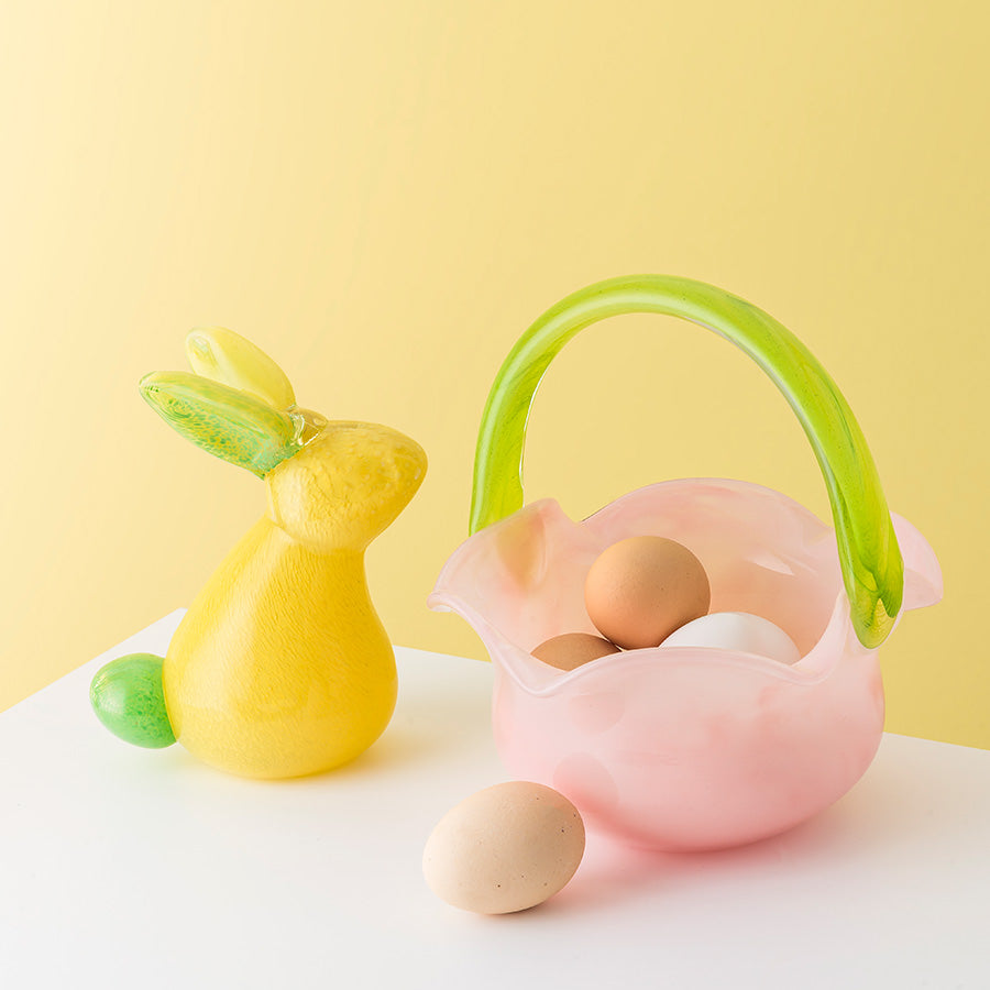 Fargeriktig kombinasjon av en rosa glasskurv med grønt håndtak og en gult påskehare, dekorert med påskeegg.