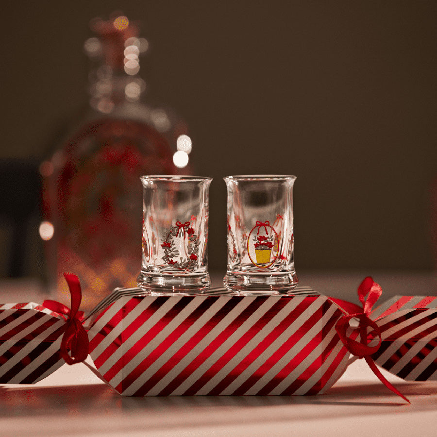 Holmegaard Christmas Juledrammeglass 2025 plassert på en julegave med røde og hvite striper, sammen med et glitrende juleservise, ideelt for festlige anledninger.