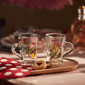 Holmegaard Christmas Hotdrink Juleglass 2025 - 23 cl, 2pk, med romantiske motiver av julekrem og røde sløyfer, perfekt for gløgg eller varm drikke, på et dekorativt brett med småkaker.