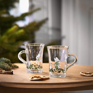 To Holmegaard Christmas 2026 hotdrink glass med juledekorasjoner på trebord, ved siden av juletre og tørkede appelsinskiver