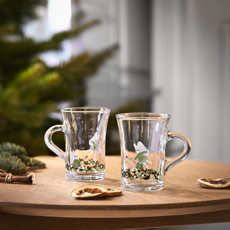To Holmegaard Christmas 2026 hotdrink glass med juledekorasjoner på trebord, ved siden av juletre og tørkede appelsinskiver