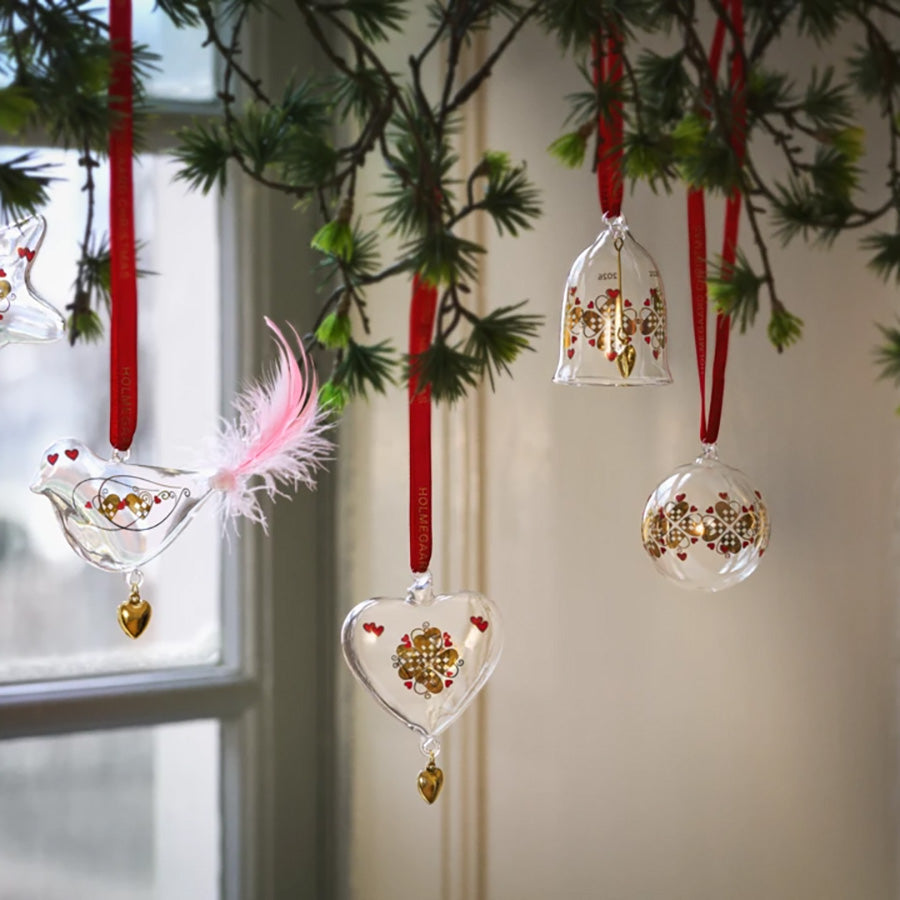 Samling av Holmegaard juleklokker og ornamenter, inkludert hjerte, fugl, og kule, alle med gylne detaljer og rød silketråd.