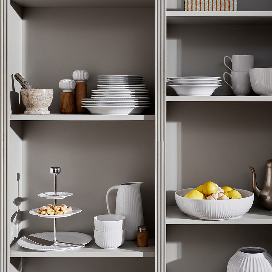 Kähler Hammershøi etasjefat 28 cm i hvit, vist på en hylle med kjøkkenutstyr og frukt, kombinerer funksjonalitet og dekorasjon.