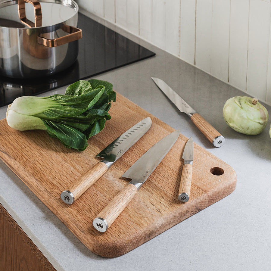 Løve Steig knivsett i bruk på et kjøkken, med fire kniver plassert på en tre skjærefjøl ved siden av grønnsaker, som viser kombinasjonen av stil og funksjonalitet.