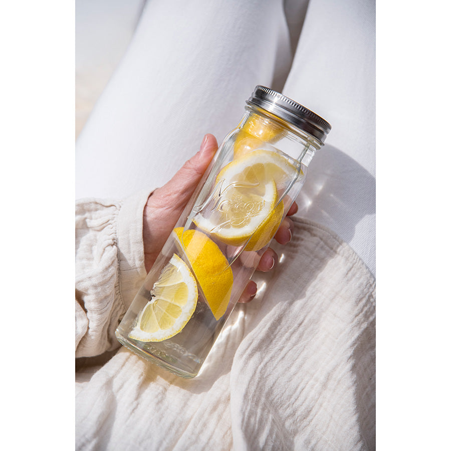 En person holder en Norgesglass flaske med skiver av sitron og iskaldt vann, perfekt for servering og oppbevaring. Flaske i klart glass med metallskrukork, ideell for hverdagsbruk.