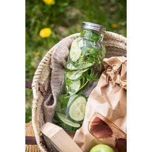 Norgesglasset flaske med agurk og mynte, liggende i en kurv med teppe og frukt, perfekt for piknik eller utendørs servering.
