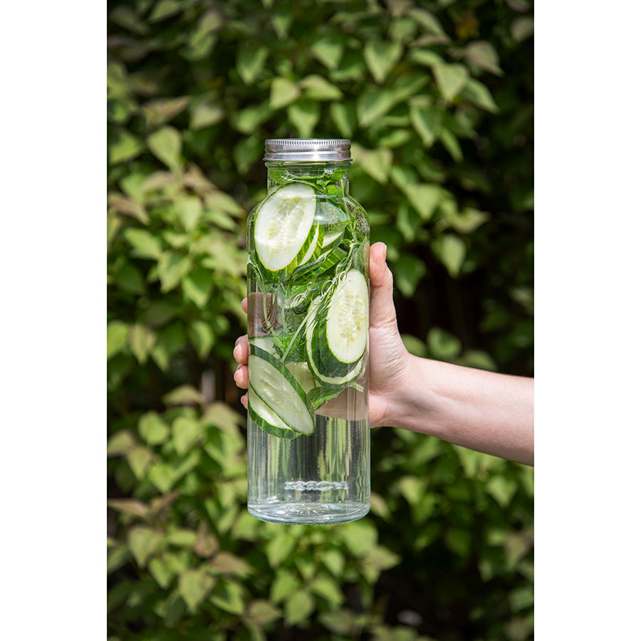 En person holder en Norgesglasset flaske med agurk og mynte, med en grønn bakgrunn, perfekt for å vise frem forfriskende drikke.