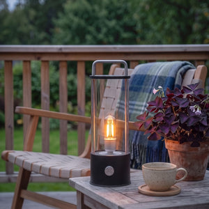 Ledlampe i sort med klart glass og bærehåndtak på terrassebord ved siden av kaffekopp