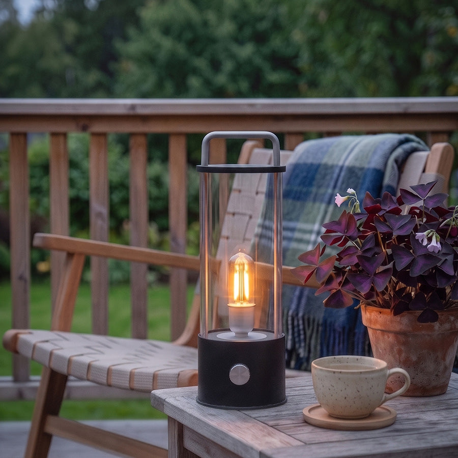 Ledlampe i sort med klart glass og bærehåndtak på terrassebord ved siden av kaffekopp