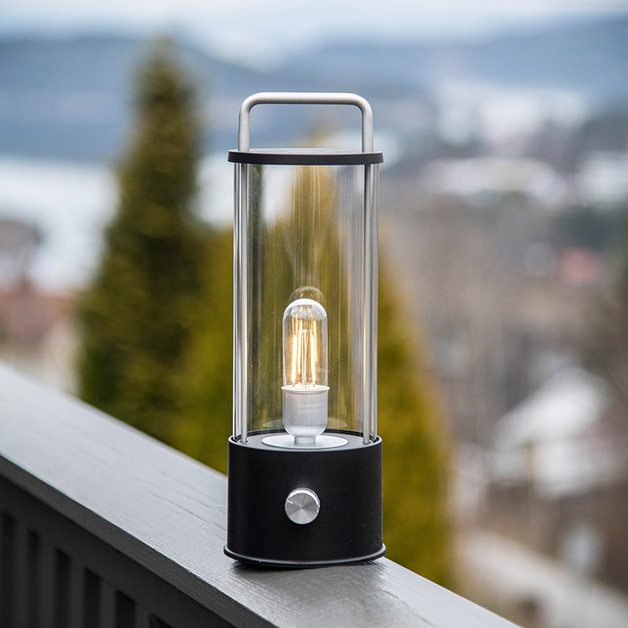 Norgesglasset Ledlampe stående på en balkong, gir et varmt lys med bakgrunn av natur og utsikt, perfekt for utendørs bruk.