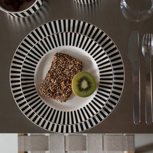 Oversiktsbilde av flere Nova Abby tallerkener med svart-hvitt stripemønster, inkludert en tallerken med kake og kiwi, på et moderne bord.