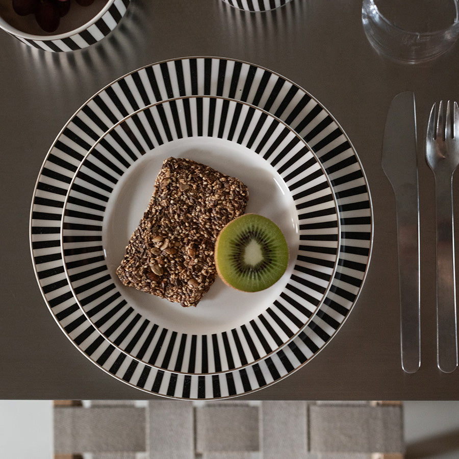 Oversiktsbilde av flere Nova Abby tallerkener med svart-hvitt stripemønster, inkludert en tallerken med kake og kiwi, på et moderne bord.