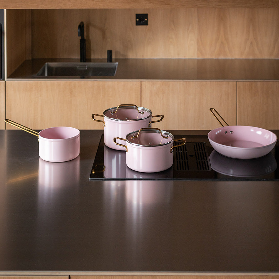 Grytesett i rosa fra Nova Faye, vist på en kjøkkenbenk med stekepanne og gryter klare til bruk med stilige gullhåndtak.