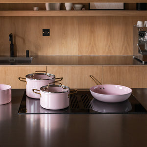 Samling av Nova Faye gryter og stekepanne på en kjøkkenbenk i tre, hvor stekepannen har en elegant rosa og gulldesign.