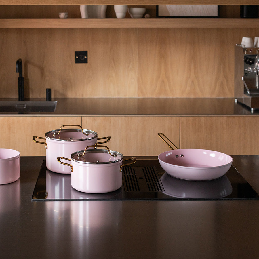 Samling av Nova Faye gryter og stekepanne på en kjøkkenbenk i tre, hvor stekepannen har en elegant rosa og gulldesign.