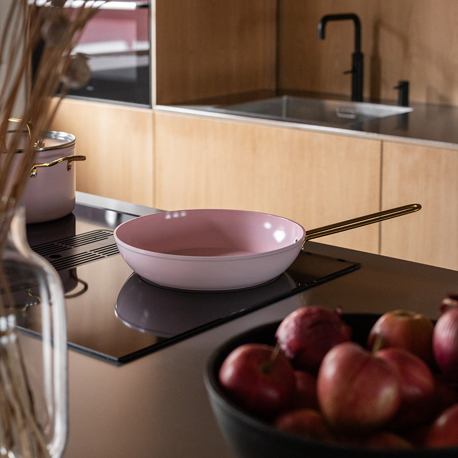 Rosa Nova Faye stekepanne med gullhåndtak på en stilren kjøkkenbenk sammen med friske epler, perfekt for innbydende måltider.