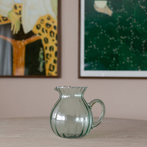 Nova Lottie mugge i optisk grønn glass, med skulpturell buet form og bølgekant, plassert på et bord med kunstverk i bakgrunnen.