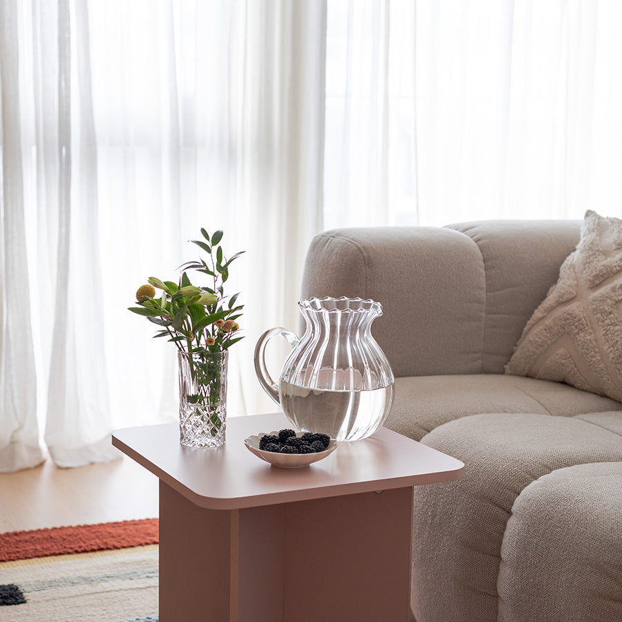 Nova Lottie mugge i klart optisk glass, plassert på et bord ved siden av en vase med blomster. Muggen har en buet silhuett og er fylt med vann, noe som viser frem det elegante designet og funksjonaliteten, perfekt for både hverdag og fest.