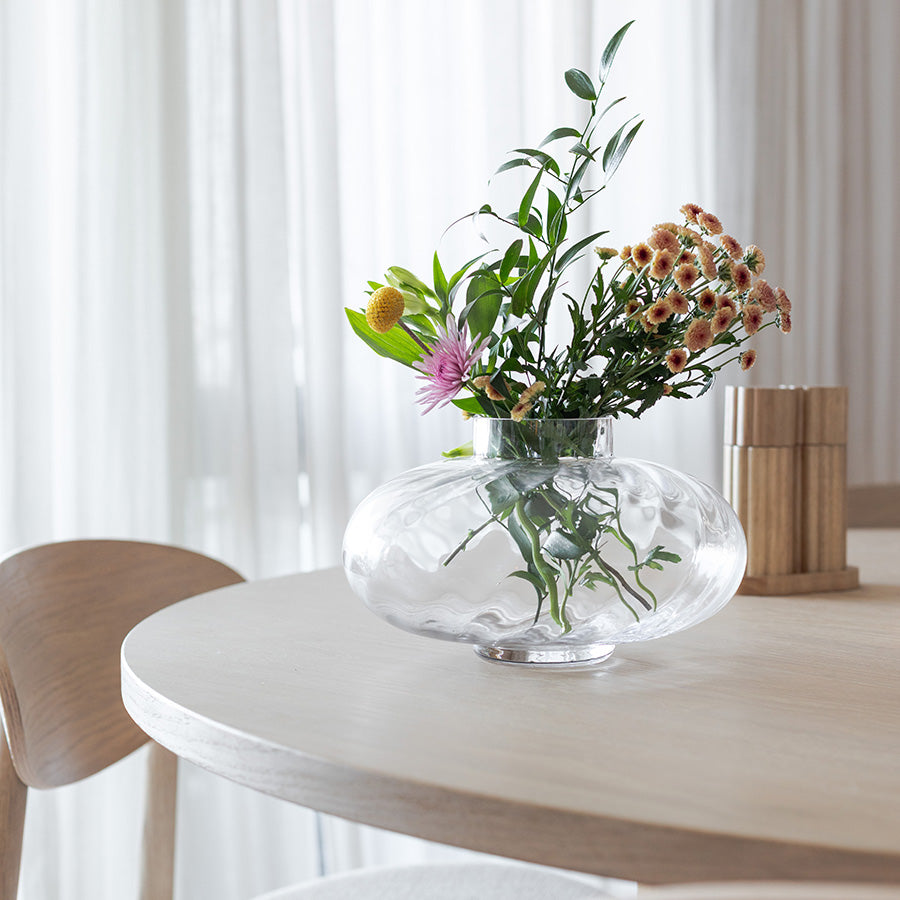 Nova Lottie vase i klart optisk glass med blomster, plassert på et lyst trebord, gir en moderne og skulpturell dekorasjon.