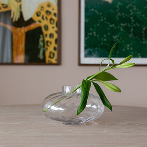Elegant Nova Lottie vase i klart glass med en grønn stilk, utstilt på et bord med kunstverk i bakgrunnen, perfekt for alle interiørstiler.