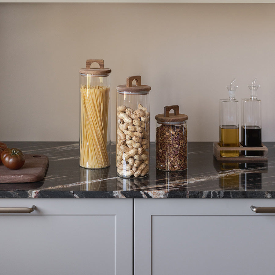 Tre oppbevaringsglass i variasjon av høyder på en moderne kjøkkenbenk. Glassene har akasietrelokk og inneholder pasta, peanøtter og tørkede blomster.