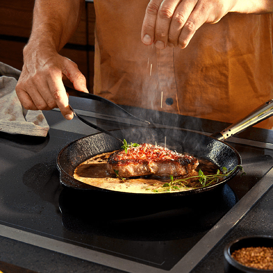 En person bruker Rosendahl Rå stekepinsett til å salte en dampende stek i en stekepanne.