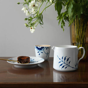 Bilde av Royal Copenhagen Blå Mega Riflet kanne 70 cl, sammen med kopp og tallerken, med fokus på den håndmalte designen og blomsterarrangementet i bakgrunnen. Perfekt for servering av melk eller juice.
