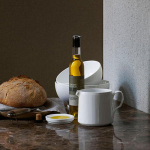 Royal Copenhagen Hvit Riflet kanne 70 cl sammen med olivenolje og ferskt brød - elegant porselenskanner som komplementerer et stilrent bordoppsett.