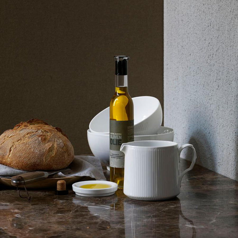 Royal Copenhagen Hvit Riflet kanne 70 cl sammen med olivenolje og ferskt brød - elegant porselenskanner som komplementerer et stilrent bordoppsett.
