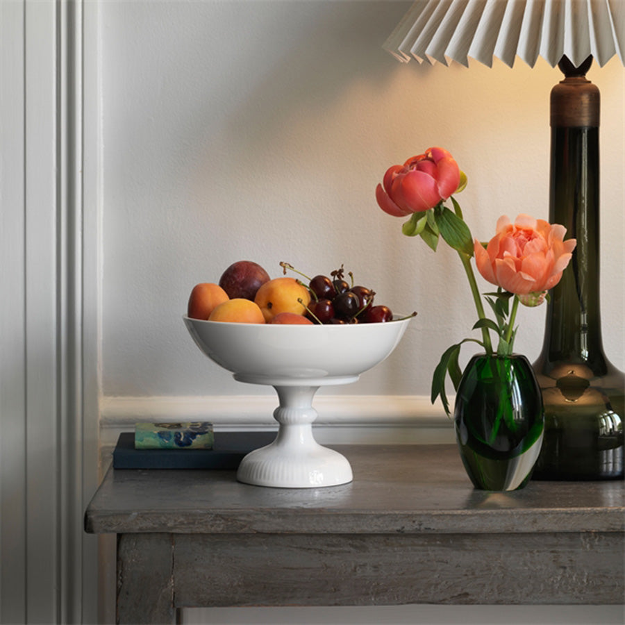 Royal Copenhagen Hvit Riflet Skål på Fot 15 cm – dekorativ hvit skål på fot med frukt, plassert på en bordflate med blomster.
