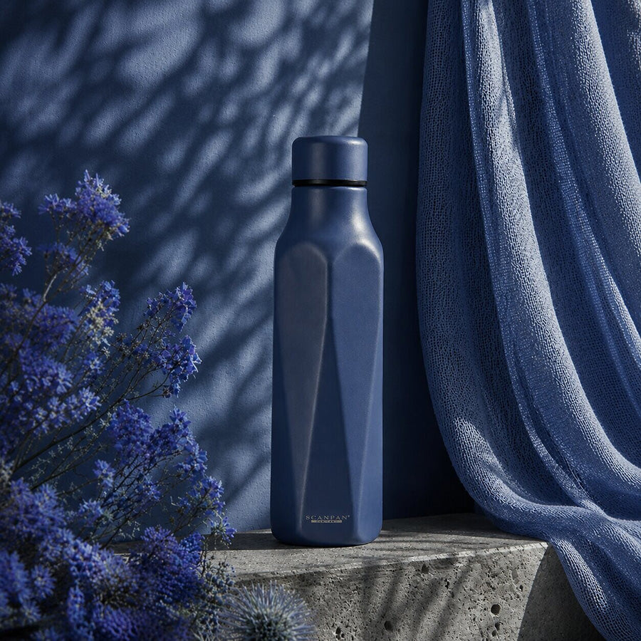 Scanpan Explore termoflaske 500 ml plassert mot en blå bakgrunn, omgitt av blomster og tekstiler som fremhever den dype oceanblå fargen og stilrene utseendet.
