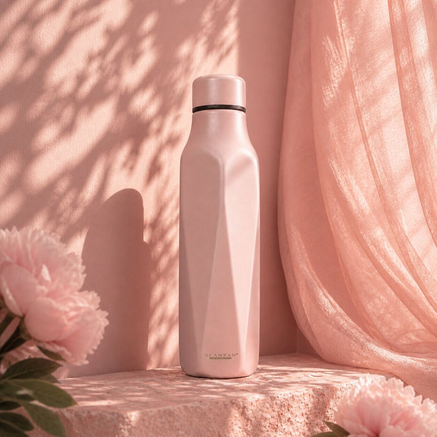 Scanpan Explore termoflaske 500 ml i rose, plassert mot en bakgrunn av myke stoffer og blomster, og fremhever dens stilige design.