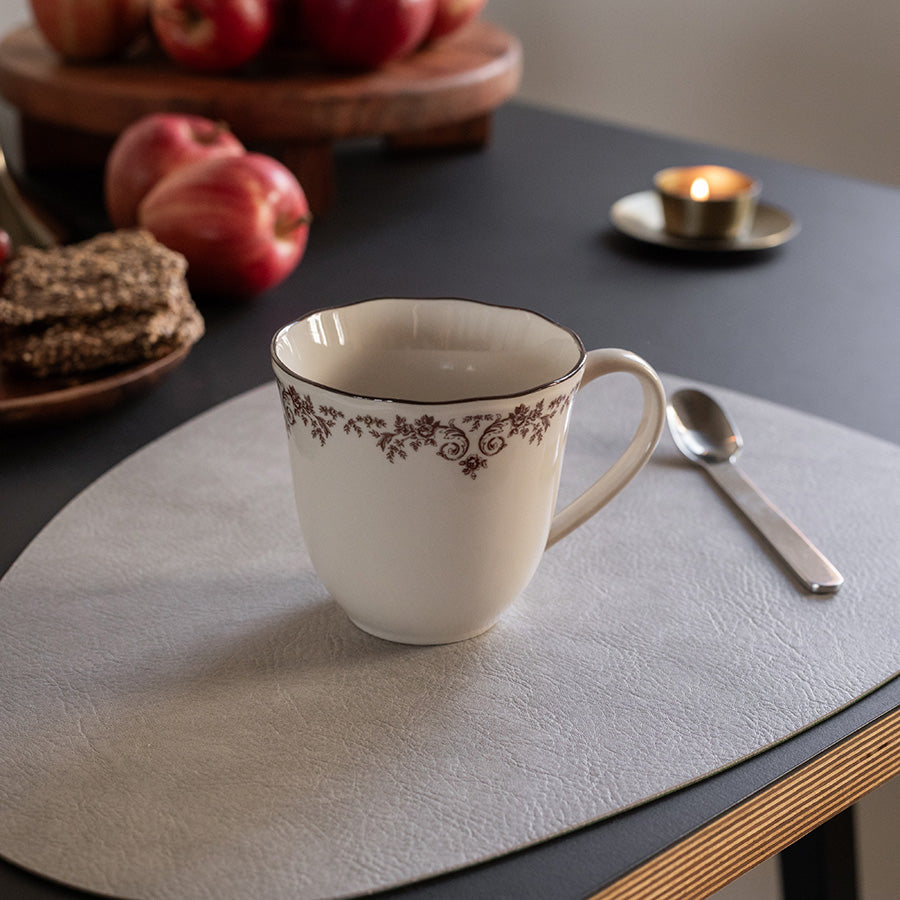 Klassisk kopp fra Seasons Fall Collection med burgunderblomstkant, satt på et moderne bord med frukt og en skje ved siden av.