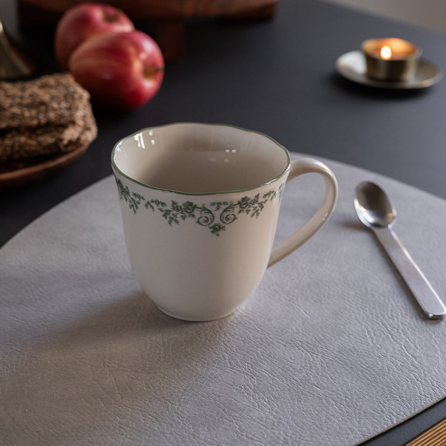 Nærbilde av Seasons Fall Collection kopp med grønn print, på et stilrent bord med epler og en skje, perfekt for melk eller te.