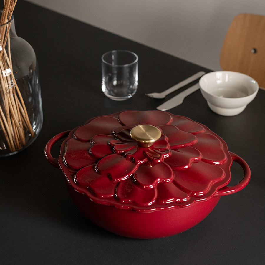 Seasons støpejernsgryte med blomsterdekor i dyp burgunderrød farge, stående på et bord med forkler, klare glass og en vase med kvister.
