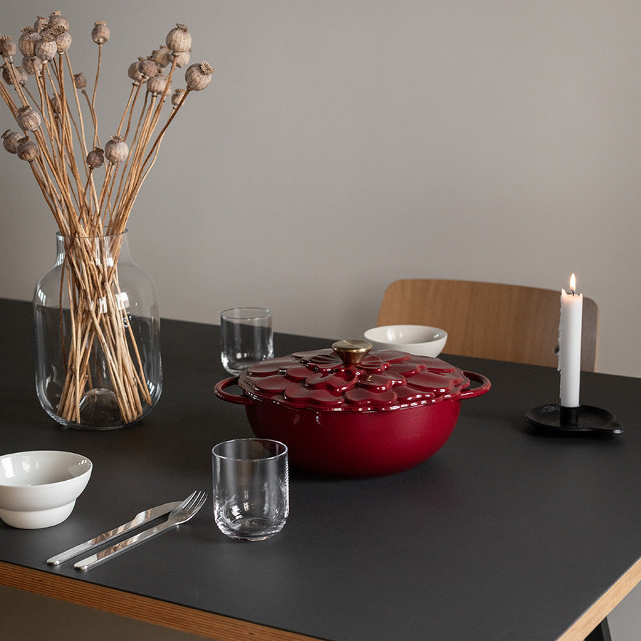 Seasons støpejernsgryte i burgunderrød med blomsterdekor, plassert på et bord med lys, glassvann og en vase med tørkede blomster, og gir en elegant middagsopplevelse.