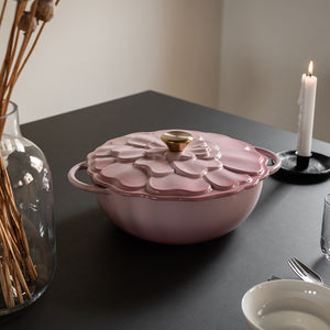 Støpejernsgryte med lokk og blomsterdesign, i en vakker rosa farge, som står på et bord, klar til bruk.