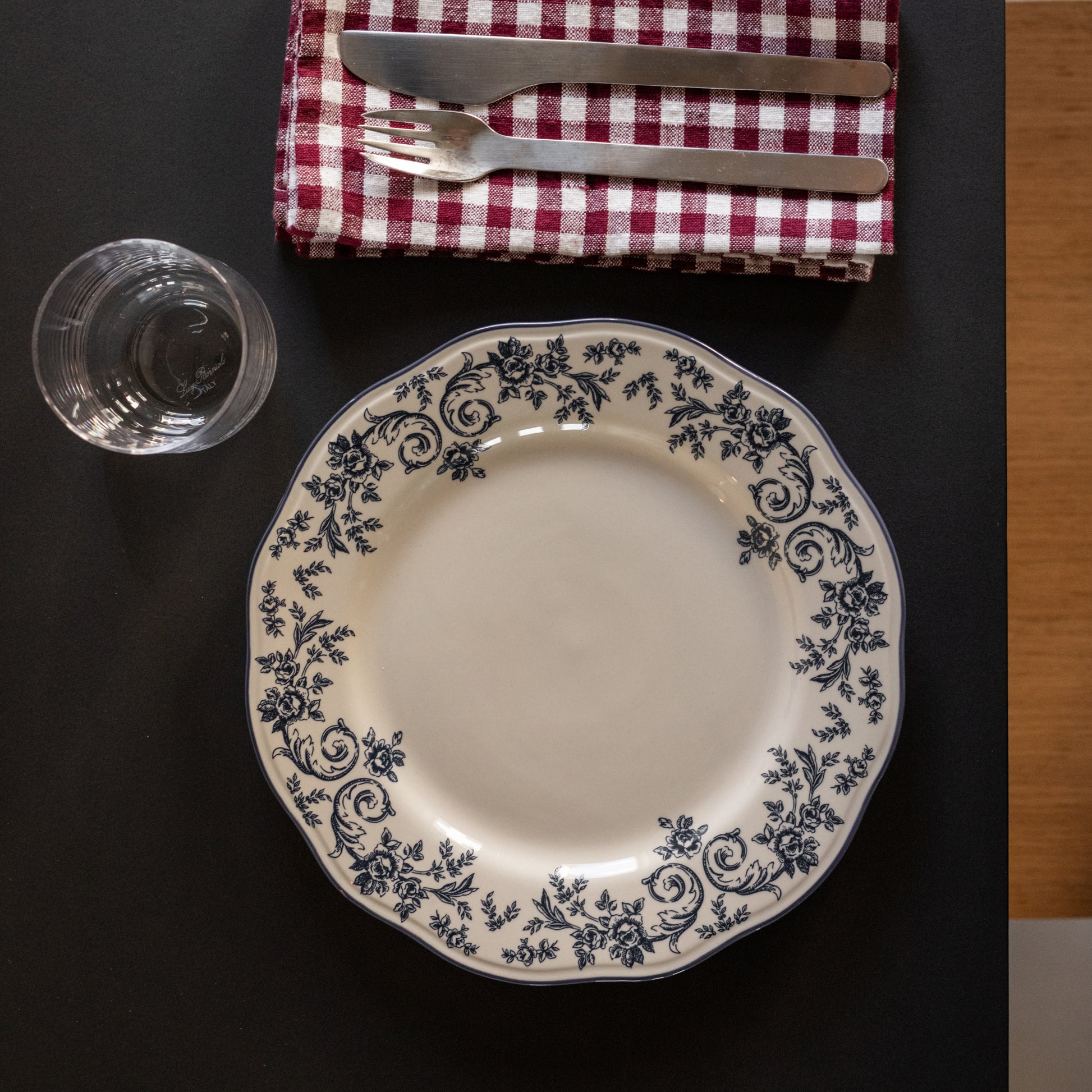 Elegant tallerken med blå blomsterprint på hvit porselen, plassert på et dekket bord med riflet rød og hvit duk, bestikk og glass.