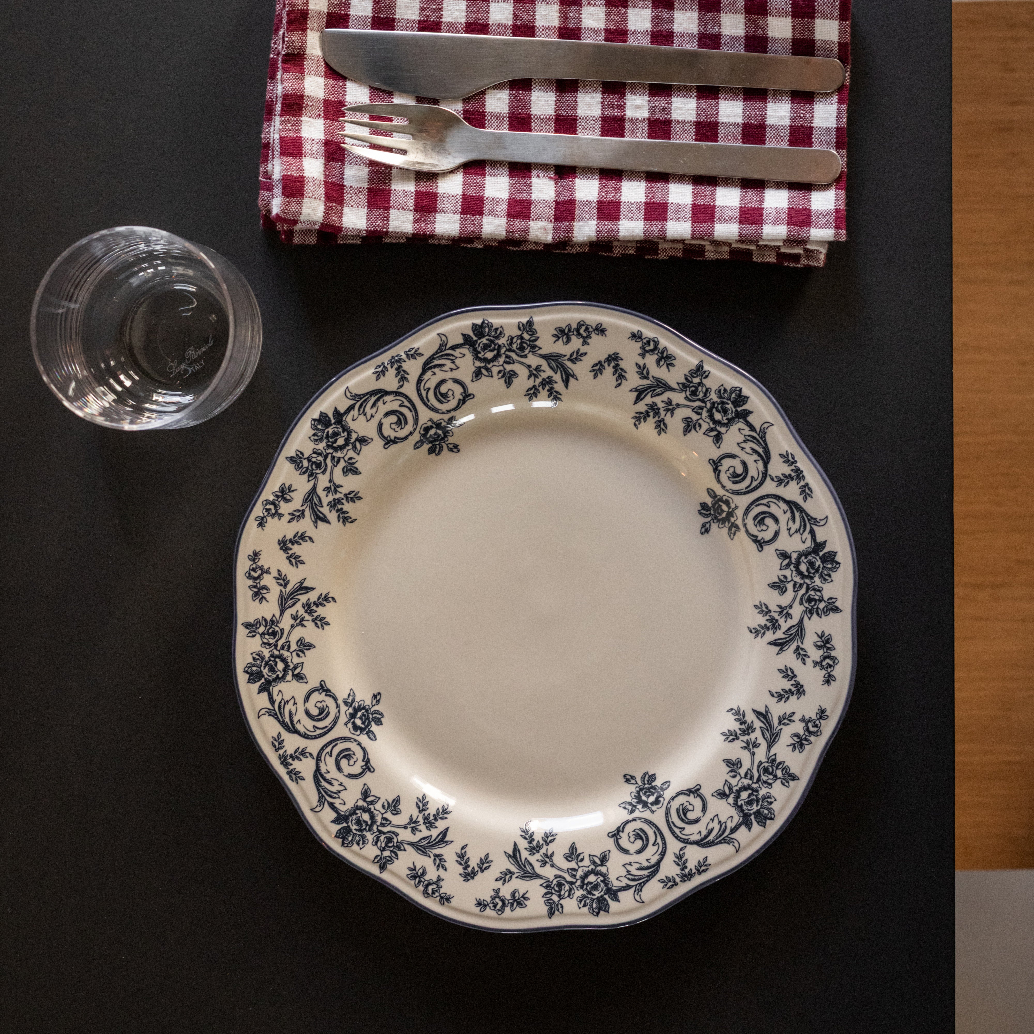 Elegant tallerken med blå blomsterprint på hvit porselen, plassert på et dekket bord med riflet rød og hvit duk, bestikk og glass.