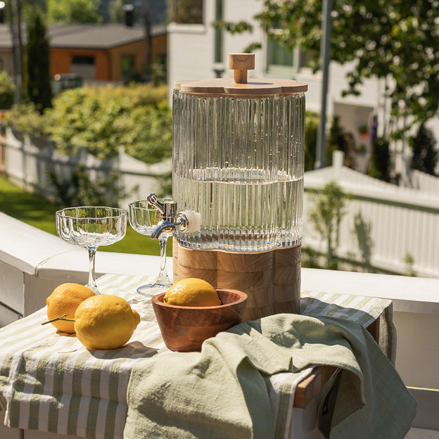 Seasons Summer Collection drikkedispenser Blomst 4L med vann og sitron, plassert på et bord i en hage, omgitt av friske sitroner og glass. Ideal for utendørsservering og sommerfester.