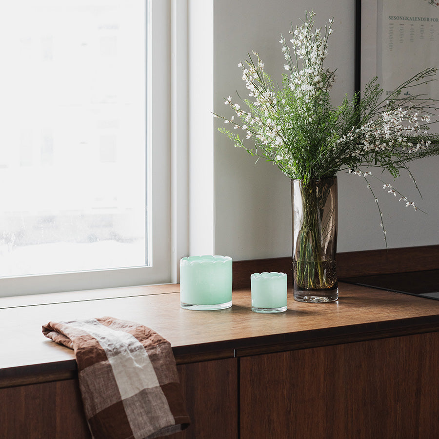 Flere lyslykter fra Seasons Spring Collection i lys grønn farge som står på en benk, sammen med en bukett av hvite blomster, skaper en varm og innbydende atmosfære.
