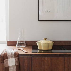 Seasons Støpejernsgryte Blomst 1.9L 30cm Gul plassert på en moderne kjøkkenbenk, sammen med en glasskaraffel og kjøkkenutstyr, demonstrerer stil og praktisk bruk.