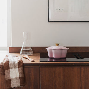 Seasons Støpejernsgryte Blomst 1,9L 30cm Rosa plassert på en moderne kjøkkenbenk med en glassvannkanne i bakgrunnen.