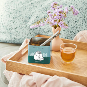 Teministeriet Moomin Te Lakris og Eple servert i en teglass med blomst i bakgrunnen og den stilige boksen på et trebrett, perfekt for en avslappende te-stund.