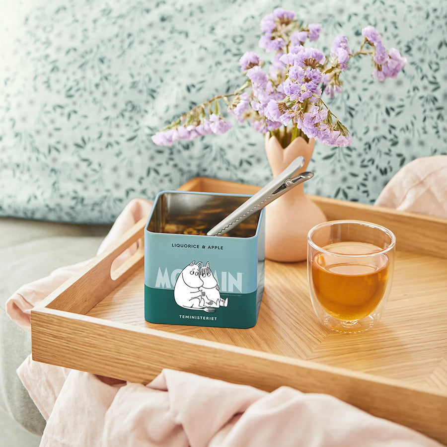 Teministeriet Moomin Te Lakris og Eple servert i en teglass med blomst i bakgrunnen og den stilige boksen på et trebrett, perfekt for en avslappende te-stund.