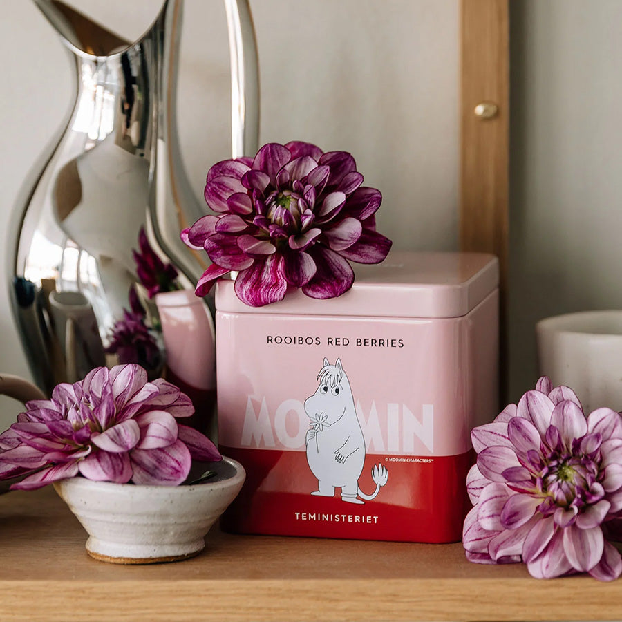 Teministeriet Moomin Rooibos Røde Bær Te pakket i en sjarmerende rosa boks, dekorert med blomster i bakgrunnen, perfekt for en fruktig og koffeinfri teopplevelse.