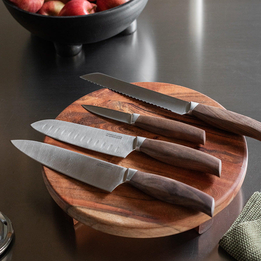 Sett med ulike kniver inkludert en santokukniv fra Christiania Glasmagasin, presentert på et trebrett.