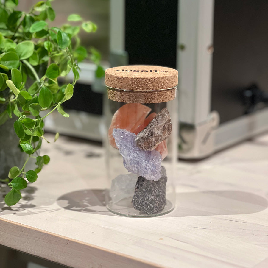 Visning av en glasstube med variasjoner av steinsalt, som Brun Østerriksk Alp salt og Boliviansk Rose salt, med korklokk, presentert i et stilrent miljø.