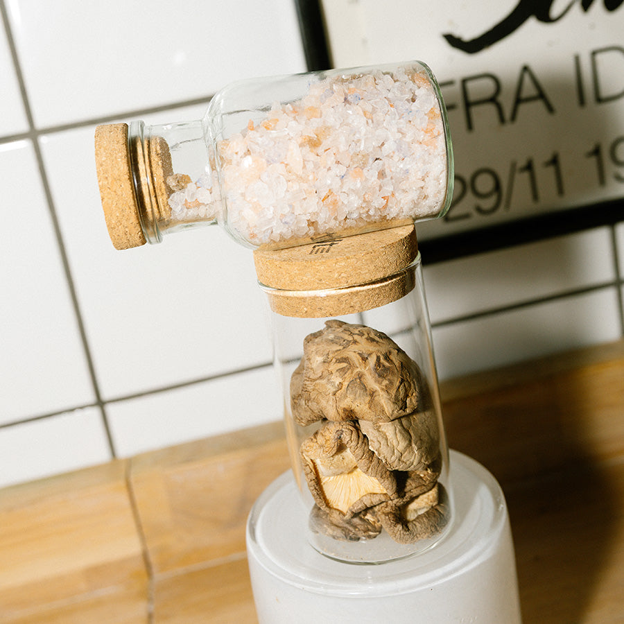 Adelante Rivsalt Rock Salt Blend i en glassbeholder med kork, plassert oppå en annen beholder med sopp. Stilig og funksjonell oppbevaring av salt.