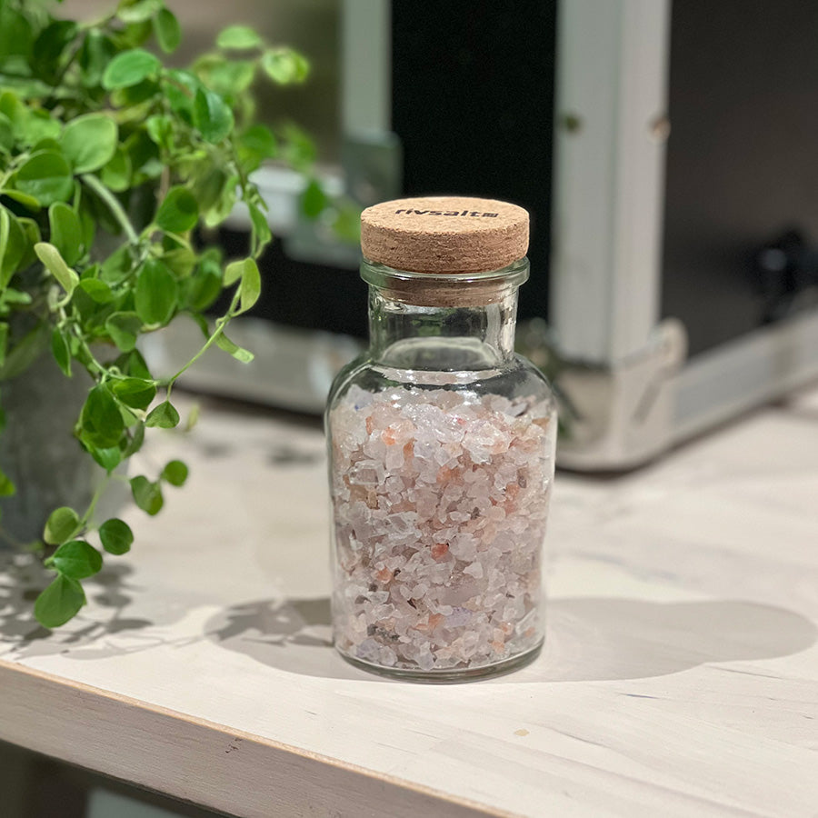 Adelante Rivsalt Rock Salt Blend i en glassflaske med kork, stående på et bord med grønn plante rundt. Ideell for å tilsette smak til hverdagsretter.
